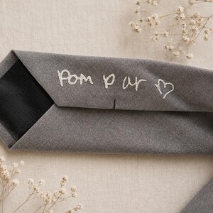 May include: A grey necktie featuring white embroidered text that reads "Pom par" followed by a small heart symbol. The tie has a black fabric lining visible on the interior fold and a smooth, matte-textured grey exterior fabric surface.
