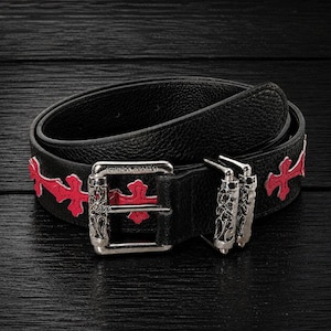 Pode incluir: Um cinto de couro preto texturizado com uma fivela de metal prateado com gravações e o texto CHROME HEARTS. A tira do cinto é decorada com uma série de aplicações em forma de cruz vermelho-brilhante posicionadas ao longo da banda.