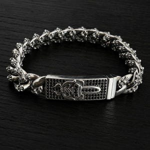May include: A silver chain link bracelet featuring a rectangular centre plate adorned with small black stones and an engraved emblem. The bracelet links are detailed with small cross motifs throughout the entire length of the silver metal band.