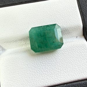 May include: A rectangular emerald gemstone with a faceted cut, displaying a deep green color with natural internal inclusions and a polished surface, positioned to showcase its geometric shape and reflective properties under bright lighting.