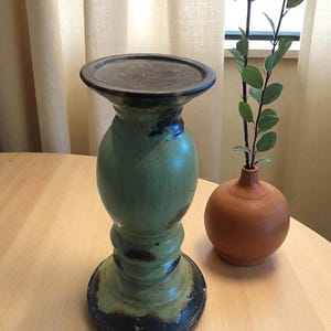 May include: A tall, cylindrical candle holder with a distressed sage green and dark brown finish, featuring a wide circular base and a flat top platform. Beside it sits a small, round, terracotta-colored vase with a narrow neck and green leaves.