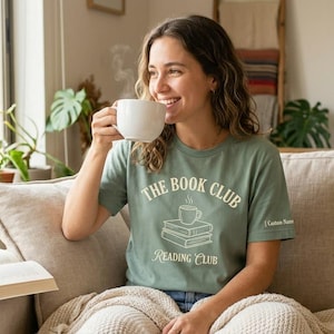 Puede incluir: Una camiseta de manga corta color verde salvia con el texto en blanco THE BOOK CLUB y READING CLUB junto a un gráfico de una taza de café sobre una pila de libros. La manga incluye el texto Custom Name en una fuente blanca pequeña.