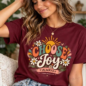 May include: A maroon short-sleeved t-shirt featuring a graphic print with the text "CHOOSE Joy ALWAYS" and "1 THESSALONIANS 5:16". The design includes a yellow sun, white daisies, green leaves, and colourful abstract shapes in orange, teal, and pink.