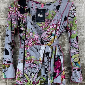 May include: A long-sleeve wrap top featuring a vibrant, multi-coloured floral and abstract pattern in shades of grey, pink, lime green, and black. The garment has a V-neckline, a side tie closure, and visible pink stitching along the seams and edges.