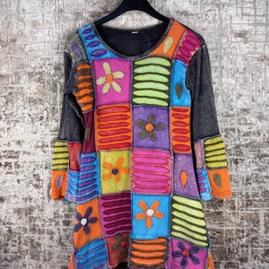 Op de afbeelding: Een tuniek met lange mouwen voorzien van een patchworkontwerp met diverse rechthoekige stoffen panelen in levendige kleuren zoals roze, oranje, blauw, paars en limoengroen. Het kledingstuk bevat gestikte bloemapplicaties en horizontale strepen.