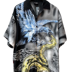 Può includere: Una camicia a maniche corte con bottoni, colletto nero e una stampa grafica di un'aquila blu che attacca un serpente giallo. La camicia presenta uno sfondo grigio e nero aerografato con motivi d'acqua sul fondo e un'etichetta nera.