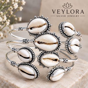 Puede incluir: Una pulsera de plata con múltiples conchas de cauri blancas engastadas en marcos de metal con cuentas ornamentadas. La pieza de joyería muestra el texto VEYLORA SILVER JEWELRY en la parte superior. El diseño incorpora varias conchas.