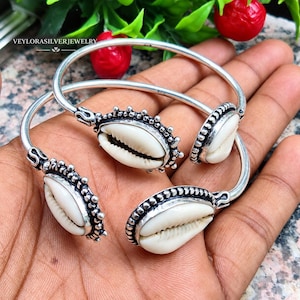 Peut inclure: Deux bracelets ouverts en argent ornés de coquillages cauris blancs sertis dans des montures perlées. Le métal présente une finition oxydée foncée dans les creux, mettant en valeur les détails texturés entourant chaque coquillage.