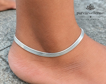 May include: A silver-toned herringbone chain anklet with a flat, flexible, and tightly woven link design. The jewelry piece features a smooth, polished surface that reflects light, creating a continuous metallic band around the ankle area.