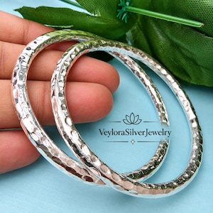 Könnte beinhalten: Zwei silberne Armreifen mit einer gehämmerten, texturierten Oberfläche, die das Licht reflektiert. Der Schmuck ist mit dem Text VeyloraSilverJewelry versehen. Die runden Armreifen haben ein poliertes Finish und eine gleichmäßige, abgerundete Dicke.