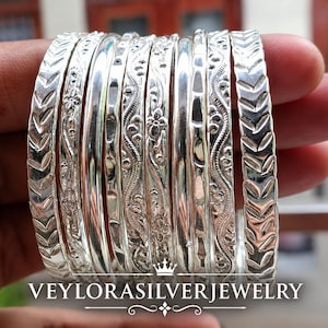 Peut inclure: Un ensemble de neuf bracelets joncs en argent présentant diverses textures et motifs, notamment des chevrons, des fleurs et des ondulations. Le texte VEYLORASILVERJEWELRY est imprimé en bas, avec une petite couronne et une ligne décorative.