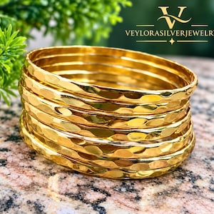 Op de afbeelding: Een stapel van zeven goudkleurige armbanden met een gehamerd, getextureerd oppervlak. Elke armband heeft een reflecterende, gepolijste afwerking die het licht opvangt. De merknaam VEYLORASILVERJEWELRY staat in witte tekst in de rechterbovenhoek.