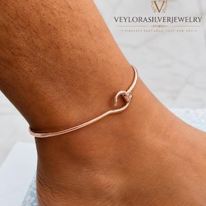 Può includere: Una cavigliera color oro rosa caratterizzata da un sottile cinturino in metallo liscio con chiusura a gancio. Il gioiello è indossato attorno alla caviglia e presenta un design essenziale con una superficie lucida, riflettente e un occhiello.