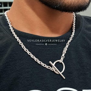 May include: A silver chain necklace featuring a toggle clasp with a circular ring and a straight bar. The necklace is worn around the neck over a black shirt. The text VEYLORASILVERJEWELRY is printed in white above the silver chain necklace.