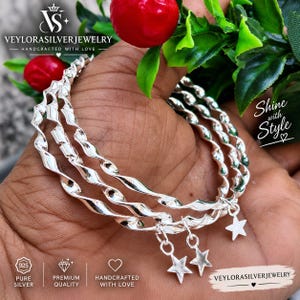 May include: A set of three twisted silver bangles featuring small dangling star charms. The bracelets are polished and reflective, showcasing a spiraled metal design. Each bangle is adorned with a single five-pointed star charm attached to the band.