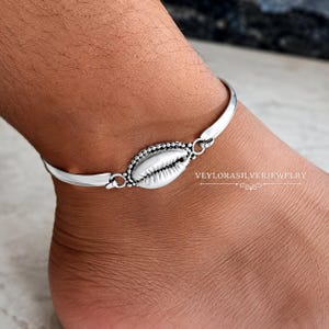 Può includere: Una cavigliera in argento con un ciondolo centrale a forma di conchiglia di ciprea con bordo perlato, collegato a una fascia in argento liscia e lucida. Il testo VEYLORASILVERJEWELRY è stampato sul lato destro dell'immagine, vicino alla cavigliera.