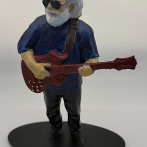 May include: A hand-painted figurine of a musician with white hair and a beard, wearing black sunglasses, a dark blue shirt, and black pants. The figure holds a red electric guitar with a brown strap, standing on a black oval base.