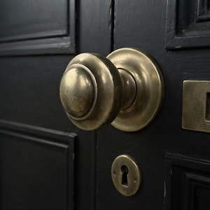 May include: A round brass door knob with a matching circular backplate, a rectangular letter slot, and a keyhole escutcheon plate mounted on a dark black paneled door. The hardware features a weathered, metallic finish with visible patina.
