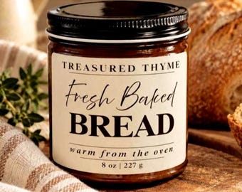 May include: A glass candle jar with a black lid and a cream-colored label. The label reads: TREASURED THYME, Fresh Baked BREAD, warm from the oven, 8 oz | 227 g. The jar contains a brown wax and is positioned upright on a wooden surface.