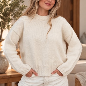 May include: A cream-coloured long-sleeve knit jumper featuring a crew neckline, dropped shoulders, and ribbed cuffs at the wrists and hem. The jumper is constructed with a medium-weight yarn and has a relaxed, slightly oversized fit throughout.
