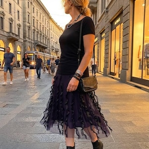 Pode incluir: Uma t-shirt preta de manga curta combinada com uma saia midi roxo-escura com camadas verticais de tule com folhos. O conjunto inclui uma mala a tiracolo preta com tachas metálicas, um colar de contas, pulseiras prateadas e botins de pele preta.