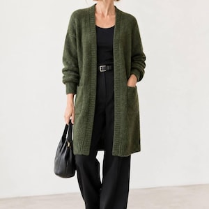 May include: An olive green open-front knit cardigan featuring long sleeves, ribbed cuffs, a ribbed hem, and two large patch pockets. The cardigan is worn over a black scoop-neck top, black trousers with a black belt, and black shoes, while carrying a black handbag.
