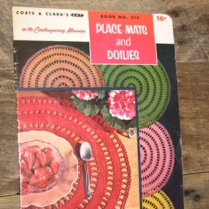 May include: A vintage book titled "Place Mats and Doilies" by Coats & Clark's ONT. The cover features colourful crochet designs in red, green, pink, and yellow. The book is open, displaying a red place mat with a plate of strawberries and a spoon.