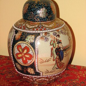 May include: A ceramic ginger jar with a lid, featuring a detailed design. The jar has a floral motif, figures in traditional dress, and a crackle glaze. The lid is dark blue with gold accents. The jar is on a red patterned surface.