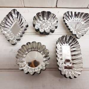 May include: Five vintage silver tartlet moulds in different shapes: diamond, flower, triangle, round, and rectangular. Each mould has a fluted edge and a shiny, reflective interior. The moulds are arranged on a white wooden surface.