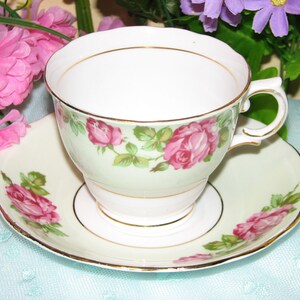 May include: A vintage teacup and saucer set. The white teacup features pink rose floral designs with green leaves and gold trim. The matching saucer has a similar design. The set is displayed on a light blue surface with pink and purple flowers in the background.