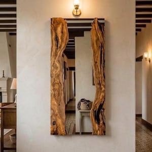 以下が含まれることがあります： 自然な木の縁取りが施された長方形の壁掛け鏡。木目模様がはっきりと現れており、側面は有機的で不規則な形状をしています。鏡は壁に垂直に取り付けられており、室内の様子と上部の小さな壁掛け照明を映し出しています。