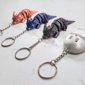 May include: Four articulated keychains featuring three dinosaur-like figures with textured, metallic-finish bodies in copper, blue, and purple-green, alongside one solid white, rounded, segmented figure, each attached to a silver metal split ring.