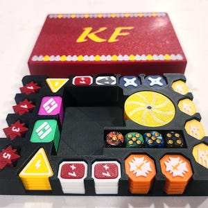 May include: A black plastic game organizer tray containing various colorful tokens, including red star-shaped pieces, yellow triangles, white squares with plus one, and orange hexagons. A red box lid with the yellow letters KF sits behind the tray.