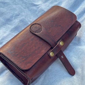 Op de afbeelding: Een donkerbruine leren oprolbare etui met een klepsluiting die wordt vastgezet met een leren riempje en twee messingkleurige metalen klinknagels. Het leer heeft een zichtbare nerf en een cirkelvormig logo op het riempje.