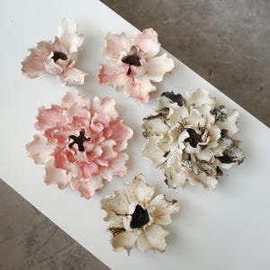 May include: Five ceramic flower sculptures with ruffled petals. Two flowers are shades of soft pink, while three flowers are off-white with dark, textured centers and black accents on the petal edges, arranged in a cluster on a flat surface.