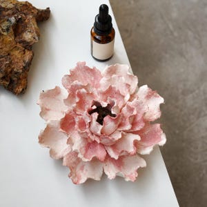 May include: A sculpted ceramic flower with layered, ruffled petals in shades of pale pink and white. The flower has a textured, matte finish. Beside it sits a small amber glass dropper bottle with a black cap and a blank white label.