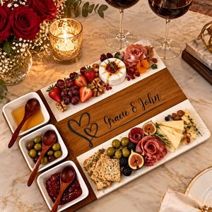 May include: A rectangular wooden and white serving board featuring the engraved text Gracie & John with a heart design. The board holds three small square white bowls with wooden spoons containing honey, olives, and jam, alongside assorted cheeses, meats, fruits, and crackers.