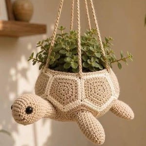 May include: A beige crochet hanging planter shaped like a turtle with a white stitched shell pattern. The turtle has black bead eyes and is suspended by four braided cords. A small green succulent plant is potted inside the turtle's shell.