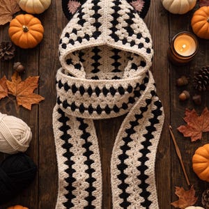 Op de afbeelding: Een gehaakte sjaal met capuchon in een crème en zwart granny square patroon met twee zwarte kattenoren bovenop. De capuchon zit vast aan een lange sjaal met bijpassende crème en zwarte stiksels, wat zorgt voor een samenhangend ontwerp.