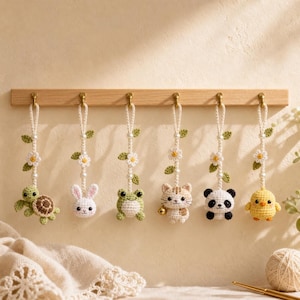May include: Six crocheted animal ornaments hanging from a wooden rack. The figures include a turtle, rabbit, frog, cat with a small bell, panda, and chick. Each ornament features a white crocheted cord decorated with small white flowers and green leaves.
