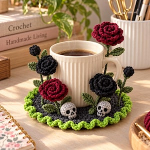 Puede incluir: Una taza de cerámica blanca se asienta sobre un posavasos redondo de ganchillo gris oscuro con un borde ondulado verde brillante. El posavasos está decorado con rosas de ganchillo rojas y negras en relieve sobre tallos verdes y dos pequeñas calaveras de ganchillo grises.