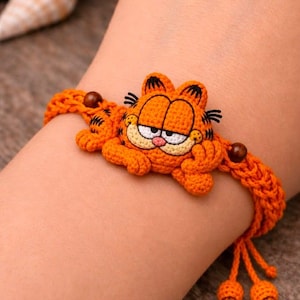 May include: A handmade orange crochet bracelet featuring a detailed Garfield character face with black stripes, white eyes, and a pink nose. The band is constructed from braided orange yarn and accented with small brown wooden beads and dangling ends.