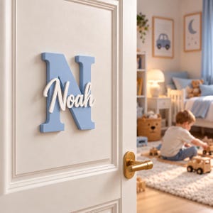 May include: A light blue wooden letter N mounted on a white door, featuring the name Noah in white cursive script layered over the letter. The sign is positioned above a brass-colored door handle on a white paneled door surface.