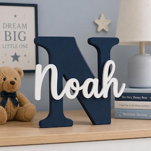 May include: A large, dark navy blue wooden letter N with the name Noah written in white cursive script across the front. The letter is freestanding and features a smooth, matte finish, serving as a decorative piece for a nursery or bedroom shelf.