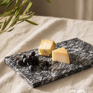 Può includere: Un tagliere rettangolare in marmo venato bianco e nero. Il tagliere sostiene un piccolo grappolo di uva viola scuro, uno spicchio di formaggio giallo pallido con venature blu e uno spicchio di formaggio sodo di colore chiaro.