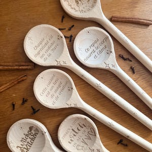 May include: Five light-coloured wooden spoons with laser-engraved text and star motifs. The spoons feature phrases including "Support Your Local Murder," "Chaque ingrédient est un cadeau de la Terre," "ce repas nourrit le corps et l'âme," "Witch Kitchen," and "There's a little witch in all of us."