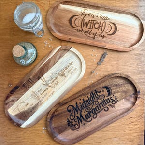 May include: Three oval wooden trays with laser-engraved text and illustrations. One tray reads 'There is a little witch in all of us' with moon phases. Another features 'Midnight Margaritas' with a crescent moon. The third lists herbal folklore.