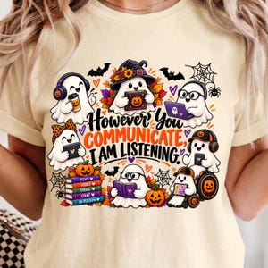 Puede incluir: Una camiseta de color crema con un gráfico de varios fantasmas de dibujos animados realizando diversas actividades. El texto dice: "However you communicate, I am listening". El diseño incluye calabazas, hojas de otoño, murciélagos y telarañas.