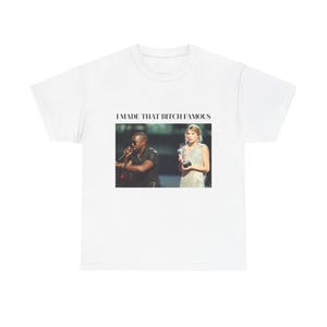 May include: A white short-sleeved t-shirt featuring a central rectangular graphic print. The image depicts a person in a black shirt holding a microphone next to a person in a silver dress holding an award. The text I MADE THAT BITCH FAMOUS is printed in black above the image.