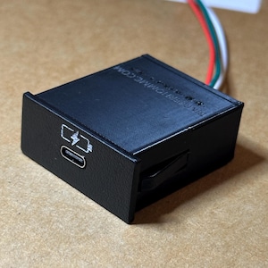 Może przedstawiać: Czarny, prostokątny elektroniczny moduł ładujący z portem USB-C na przedniej ściance oraz nadrukowaną białą ikoną baterii i błyskawicy. Urządzenie posiada teksturowane wykończenie oraz czerwone, zielone i białe przewody wychodzące z tyłu obudowy.